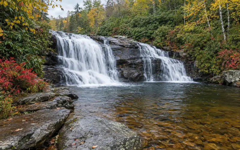 Lower Falls tumbling over a dark rock ledge into a clear pool at Graveyard Fields, framed by rhododendron and early fall color
